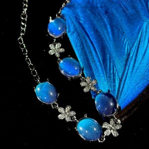 Könnte beinhalten: Eine silberne Halskette mit ovalen, blauen Edelsteinen und kleinen Blumenakzenten. Die Halskette ist vor einem leuchtend blauen Hintergrund mit schwarzem Hintergrund platziert. Die Edelsteine haben eine schimmernde, irisierende Qualität.