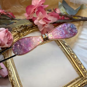 May include: A decorative hair barrette with a bow-tie design. The barrette features a gold-colored metal frame and is filled with iridescent, pink, and purple glittery material. The barrette is approximately 4 inches long.