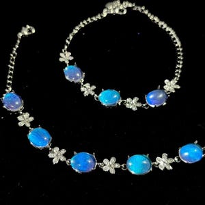Könnte beinhalten: Ein silberfarbenes Schmuckset mit Halskette und Armband. Beide Teile haben ovale, schillernde blaue Edelsteine und kleine, blütenförmige Akzente. Der Schmuck ist vor einem schwarzen Hintergrund platziert.