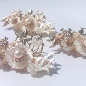 Könnte beinhalten: Drei verzierte Haarspangen mit floralen und Perlenakzenten. Die Spangen haben goldfarbenes Metall, weiße Blüten und Kunstperlen. Das Design umfasst zarte Blätter und eine korallenartige Struktur, die eine elegante Ästhetik erzeugt.