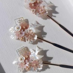 Könnte beinhalten: Drei dekorative Haarnadeln mit Blumen- und Vogelmotiven. Jede Nadel hat eine Ansammlung von rosa und weißen Blumen, schillernden Blättern und einem kleinen Vogel. Die Stiele der Nadeln sind goldfarben und auf einer weißen Oberfläche angeordnet.