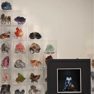 Könnte beinhalten: Eine Sammlung verschiedener Mineralienproben in einzelnen, klaren Acrylwürfeln, die treppenförmig angeordnet sind, neben einer dunkelblauen Mineralienprobe in einem quadratischen schwarzen Rahmen mit Innenbeleuchtung.
