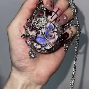 Könnte beinhalten: Silberner Taschenuhr-Anhänger mit Kette. Das Zifferblatt der Uhr zeigt ein blaues Schmetterlingsdesign und römische Ziffern. Die Uhr ist mit rosa floralen Akzenten und einem kleinen Glockenanhänger verziert. Die Kette ist ein filigranes, verbundenes Design.