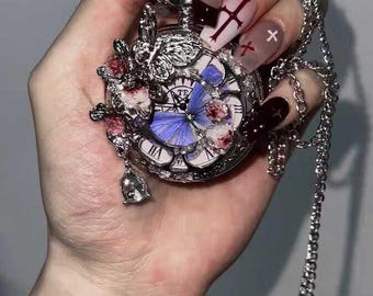 May include: Silver pocket watch pendant with a chain. The watch face features a blue butterfly design and Roman numerals. The watch is adorned with pink floral accents and a small bell charm. The chain is a delicate, linked design.