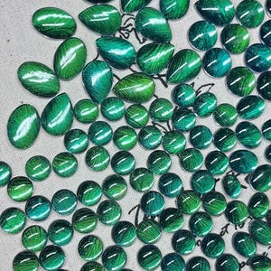 Könnte beinhalten: Eine Auswahl an grünen, ovalen und runden Glascabochons mit einer strukturierten, irisierenden Oberfläche. Die Cabochons sind in verschiedenen Größen erhältlich und auf einer neutralfarbenen Oberfläche angeordnet. Die Cabochons haben einen Katzenaugeneffekt.
