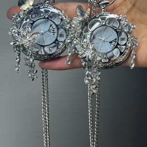 Könnte beinhalten: Zwei silberfarbene Taschenuhren mit aufwendigen Blumen- und Schmetterlingsverzierungen. Die Zifferblätter zeigen ein hellblaues Schmetterlingsdesign. Jede Uhr hängt an einer langen, filigranen Kette mit hängenden Kristallakzenten.