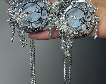 May include: Two silver-toned pocket watches with intricate floral and butterfly embellishments. The watch faces feature a light blue butterfly design. Each watch hangs from a long, delicate chain, with dangling crystal accents.