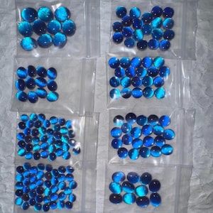 May include: Multiple clear plastic bags filled with blue cabochon gemstones. The gemstones have a cat's eye effect, with a bright blue color and a reflective quality. The bags are arranged in a grid pattern.