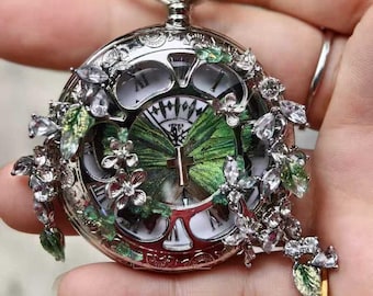 May include: A silver pocket watch with a butterfly design on the face. The watch is adorned with clear crystals and green and silver floral accents. The watch is held in a hand.