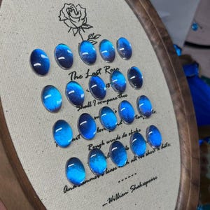 Könnte beinhalten: Ein Stickrahmen mit Leinenstoff, der eine Rosenillustration und den Text "The Last Rose" sowie ein Zitat von William Shakespeare zeigt. Ovale, blaue Cabochons sind auf dem Stoff angeordnet.
