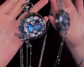 Könnte beinhalten: Zwei silberne Taschenuhren mit langen Ketten. Eine Uhr hat ein blaues Schmetterlingsdesign auf dem Zifferblatt und blaue Blumenakzente. Die andere Uhr hat ein dekoratives blaues Blumendesign. Beide Uhren werden vor einem dunklen Hintergrund gehalten.