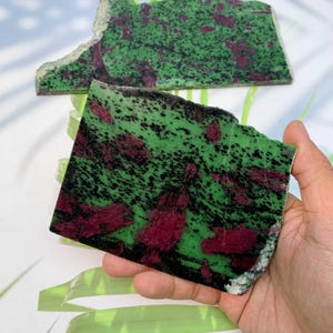 Könnte beinhalten: Zwei grob geschnittene Platten aus Rubin in Zoisit. Die Platten haben eine grüne Basis mit schwarzen Sprenkeln und großen, unregelmäßigen Flecken in tiefem Rot. Die Kanten sind uneben, und eine Platte wird in einer Hand gehalten.