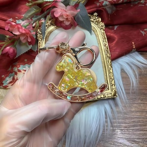 Könnte beinhalten: Ein goldfarbener Schlüsselanhänger mit einem Schaukelpferd-Design. Das Pferd ist gelb mit schillernden Akzenten, und die Basis hat rosa und rote Details. Der Schlüsselanhänger hat einen Karabinerverschluss zur einfachen Befestigung.