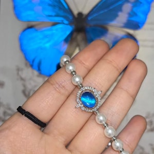 Könnte beinhalten: Ein filigranes Armband mit einem zentralen ovalen blauen Edelstein, umgeben von kleinen klaren Steinen, und verbunden mit einer Reihe weißer Perlen. Das Armband wird vor einem blauen Schmetterling präsentiert.