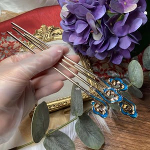 Op de afbeelding: Een set goudkleurige haarstokjes met blauwe en gouden emaille wolkaccenten. De haaraccessoires worden in een gehandschoende hand gehouden, met paarse bloemen en een decoratief gouden frame op de achtergrond.