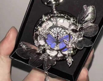 May include: Silver pocket watch with butterfly embellishments. The watch face is white with black Roman numerals and a blue butterfly design in the center. The watch is presented in a black box.