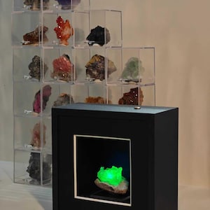 Könnte beinhalten: Eine schwarze, quadratische Vitrine mit einer internen Beleuchtung, die ein leuchtend grünes Mineralexemplar erhellt. Dahinter befindet sich eine gestufte Anordnung aus transparenten Acrylwürfeln mit verschiedenen bunten Mineralien unterschiedlicher Form und Größe.