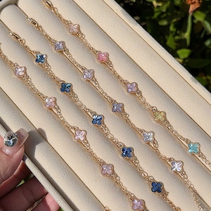 May include: Four gold-tone metal chain bracelets featuring multiple four-leaf clover charms. Each clover charm is inlaid with a small, coloured stone in various shades including pink, blue, white, and yellow. The bracelets have a lobster claw clasp.