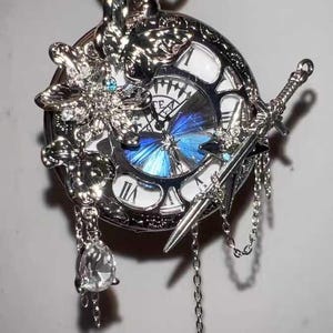Könnte beinhalten: Silberne Taschenuhr mit detailliertem Design. Die Uhr hat römische Ziffern, ein blaues Schmetterlingsdesign auf dem Zifferblatt und dekorative Elemente, darunter ein Schwert und ein Kristallanhänger. Die Uhr hängt an einer silbernen Kette.