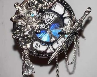May include: Silver pocket watch with a detailed design. The watch features Roman numerals, a blue butterfly design on the face, and decorative elements including a sword and a crystal pendant. The watch is suspended from a silver chain.