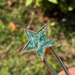 Könnte beinhalten: Eine goldfarbene Haarspange mit einem sternförmigen Ornament. Der Stern hat einen perlenartigen Rand und ist mit irisierenden, mosaikartigen Stücken in Blau-, Grün- und Türkistönen gefüllt, die das Licht mit einem Perlmutteffekt reflektieren.