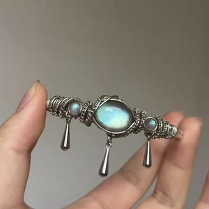 Könnte beinhalten: Ein silberfarbenes, drahtgewickeltes Armband mit einem großen, ovalen Labradorit-Stein in der Mitte und zwei kleineren, runden Labradorit-Steinen. Drei glatte, polierte, silberfarbene Metallanhänger in Tropfenform hängen an der Drahtfassung.