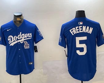 May include: Two blue baseball jerseys. One jersey has the text "Dodgers" in white with a white LA logo. The other jersey has the text "Freeman" in white with the number "5" in white.