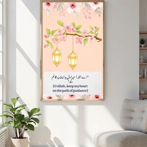 Puede incluir: Una impresión enmarcada con una ilustración en acuarela. La obra de arte representa una rama con flores rosas y linternas colgantes sobre un fondo melocotón. La impresión incluye texto en árabe e inglés: "O Allah, keep my heart on the path of guidance."