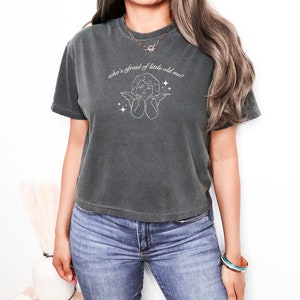 Peut inclure: Un t-shirt court gris foncé avec un contour blanc d'un ange avec des ailes et le texte "who's afraid of little old me?"