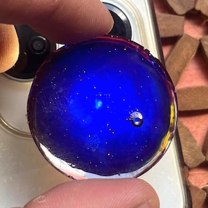 Può includere: Un supporto circolare per telefono in resina traslucida, di colore blu intenso con particelle di glitter dorate incorporate e una piccola bolla d'aria visibile sulla superficie. L'oggetto è posizionato sopra il retro di uno smartphone chiaro.