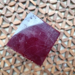 Può includere: Un blocco quadrato di colore bordeaux intenso con una superficie liscia e riflettente. L'oggetto presenta lievi graffi visibili sulla faccia superiore e bordi netti e definiti, offrendo un aspetto solido e uniforme di colore rosso scuro.