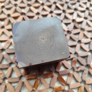 Può includere: Un blocco quadrato di sapone marrone scuro con un visibile motivo circolare a venature di legno sulla superficie superiore. Il sapone ha una consistenza liscia e opaca e un aspetto solido e uniforme, caratterizzato da un colore terra profondo.