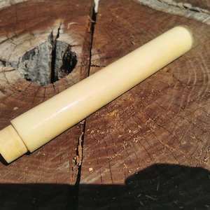 Può includere: Un oggetto cilindrico in legno bianco sporco, con un'estremità arrotondata e un tappo rimovibile di colore più chiaro. È appoggiato su una superficie di legno marrone usurata con venature e nodi visibili. L'oggetto sembra essere un contenitore o uno strumento.