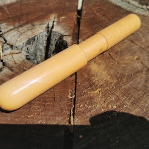 Op de afbeelding: Een lichtbruin, cilindrisch object met een afgerond uiteinde en een licht gestructureerd oppervlak. Het object lijkt gemaakt te zijn van hout of een vergelijkbaar materiaal. Het ligt op een verweerd, bruin houten oppervlak.