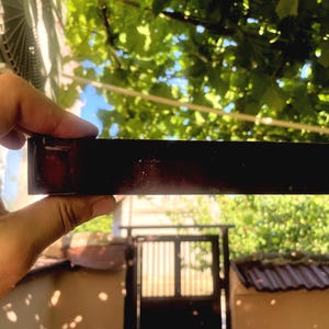 Puede incluir: Un bloque rectangular con un acabado oscuro, rojizo y translúcido. El objeto tiene una superficie lisa y pulida que capta la luz, revelando tonos profundos de ámbar y caoba a lo largo de su estructura sólida, alargada y plana.