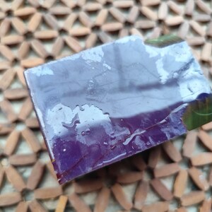 以下が含まれることがあります： 滑らかで光沢のある表面を持つ長方形の紫色の石鹸。石鹸は深い紫色をしており、縁に沿って微妙な濃い緑色の内包物が見え、表面は小さな水滴でわずかに湿っているように見えます。