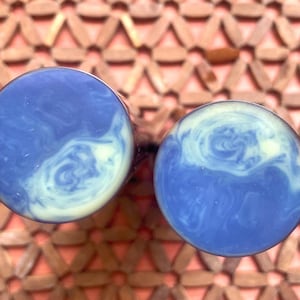 Puede incluir: Un par de expansores de oreja circulares con una superficie lisa y pulida que presenta un patrón de mármol con remolinos en colores azul profundo y blanco crema. Los expansores tienen una cara plana y redonda y una forma cilíndrica.