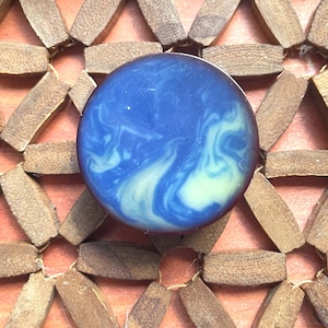 Puede incluir: Un botón circular que presenta una superficie lisa y pulida con un patrón jaspeado de remolinos en azul marino profundo y blanco crema. Los bordes del botón son de un tono marrón oscuro sólido, creando un contraste marcado con el centro azul.