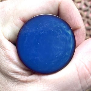 Puede incluir: Un objeto circular y plano con una superficie lisa y brillante en un color azul profundo y saturado. El centro presenta sutiles patrones de remolinos azul claro y diminutas motas blancas incrustadas en el material, creando un efecto marmolado.