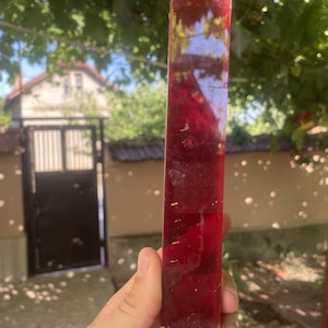 Puede incluir: Un bloque rectangular de resina roja profunda y translúcida con escamas metálicas doradas incrustadas. El objeto tiene una superficie lisa y pulida con orientación vertical, captando la luz para revelar su profundidad interna.