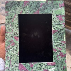 Può includere: Cornice fotografica rettangolare con bordo in stile mosaico. Il bordo della cornice presenta un motivo di frammenti di pietra verdi e rosa. Il centro della cornice è nero, progettato per contenere una fotografia. La cornice è tenuta in mano.