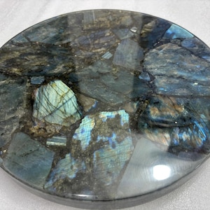May include: A circular labradorite stone with a polished surface, displaying a mosaic of blues, greens, and greys. The stone has a glossy finish and is likely used for decorative purposes or as a coaster.