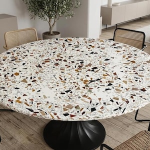 Könnte beinhalten: Runder Esstisch mit weißer Terrazzo-Oberfläche mit braunen, schwarzen und grauen Chips. Der Tisch hat einen schwarzen Sockel und ist von Stühlen mit gewebten Rücken umgeben.
