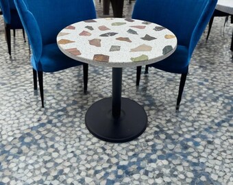 Puede incluir: Una pequeña mesa redonda con una tapa de terrazo con varios colores. La mesa tiene una base de pedestal negro y está rodeada por dos sillas de terciopelo azul. El suelo tiene un patrón de baldosas de mosaico.