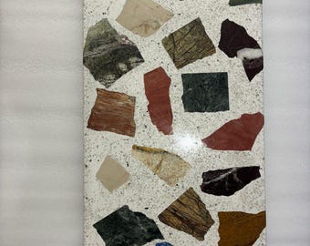 Pode incluir: Um close-up de uma superfície de terrazzo branca com várias peças de pedra coloridas e estampadas embutidas nela. A superfície é lisa e polida, com um leve brilho.