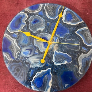 May include: Round wall clock with a blue, white, and gray agate stone design. The clock face features a unique pattern with yellow clock hands. A decorative piece for home decor.