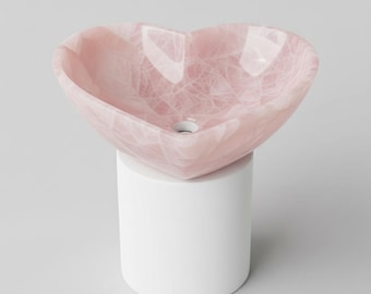 Puede incluir: Un lavabo de piedra rosa en forma de corazón sobre un pedestal blanco. El lavabo tiene una superficie pulida con vetas blancas. Un accesorio de baño único.