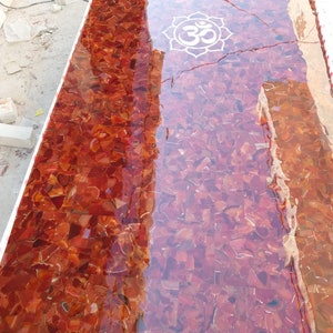 Puede incluir: Una encimera de mesa de ágata roja con un acabado de resina epoxi transparente. La encimera de mesa presenta un símbolo Om blanco en el centro.
