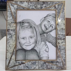 May include: A brass picture frame with a white and gray marble-like inlay. The frame is decorated with a geometric pattern. The frame is holding a black and white drawing of two children. The drawing is signed "GANGA HANDICRAFT".