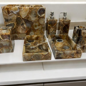 Pode incluir: Um conjunto de banheiro feito de pedra de ágata nas cores marrom, branco e cinza. O conjunto inclui um dispensador de sabonete, um porta-escovas de dentes, uma caixa de lenços de papel e duas bandejas.